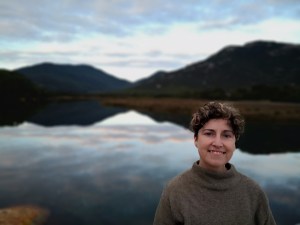 Mel, a white person with short dark brown hair wears a brown jumper and faces the camera smiling. They are to the right of the image. There are hills reflecting in a tidal pool at dusk in the background. This is blurred.